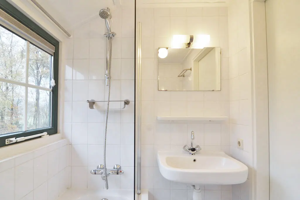 Badkamer met witte tegels, een bad met douchestang en handdouche, een glazen douchewand, een wastafel met spiegel en planchet, en een raam met een donkergroen kozijn.