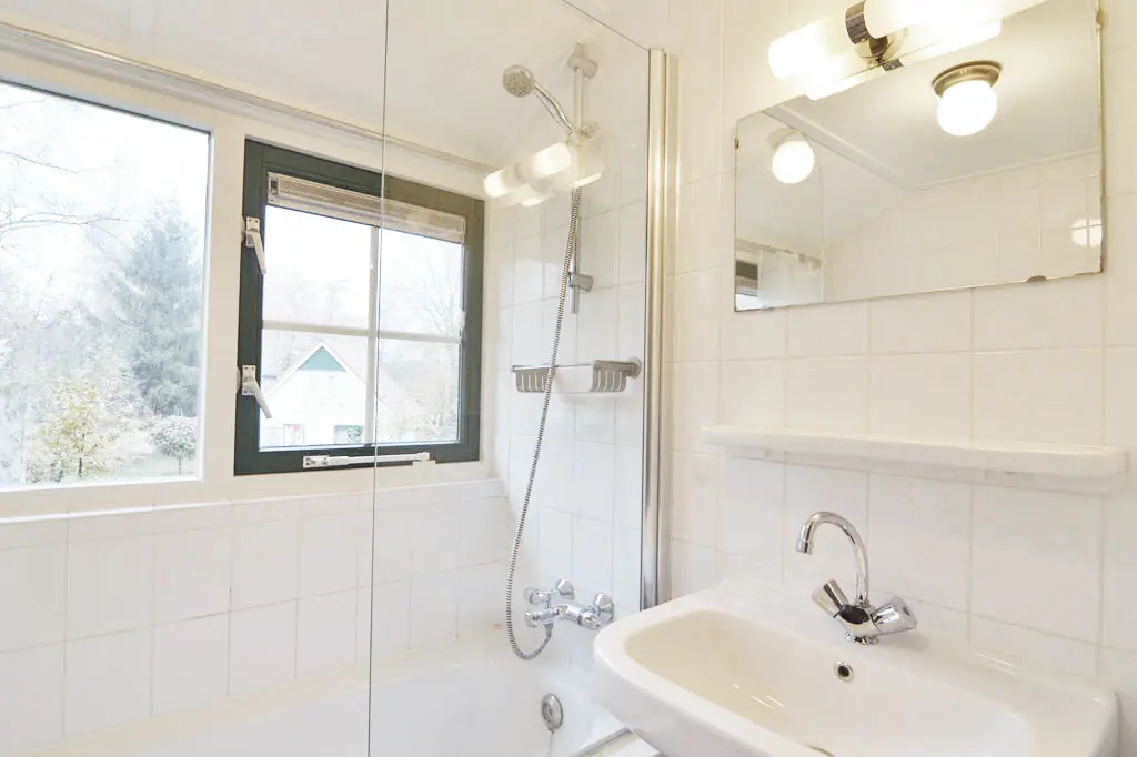Badkamer met witte tegels, een bad met handdouche en een glazen douchewand, een wastafel met spiegel en planchet, en een raam met een donkergroen kozijn.