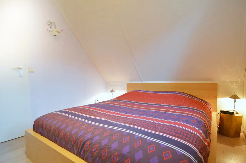 Slaapkamer onder een schuin dak met een tweepersoonsbed van licht hout, een gestreept dekbed, twee wandlampjes en een boomstamtafeltje met een wekkerradio.