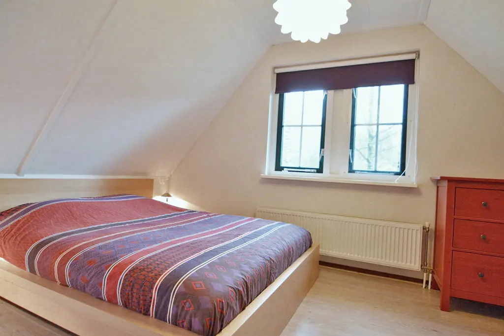 Slaapkamer met schuin dak, een tweepersoonsbed van licht hout met gestreepte sprei, een raam met een donker rolgordijn, een radiator eronder en een rode ladekast met drie lades.