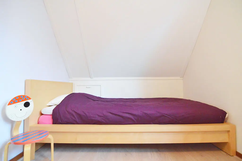 Slaapkamer onder een schuin dak met een eenpersoonsbed van licht hout, een paars dekbed en een kinderstoeltje met een geschilderd piratengezicht op de rugleuning.