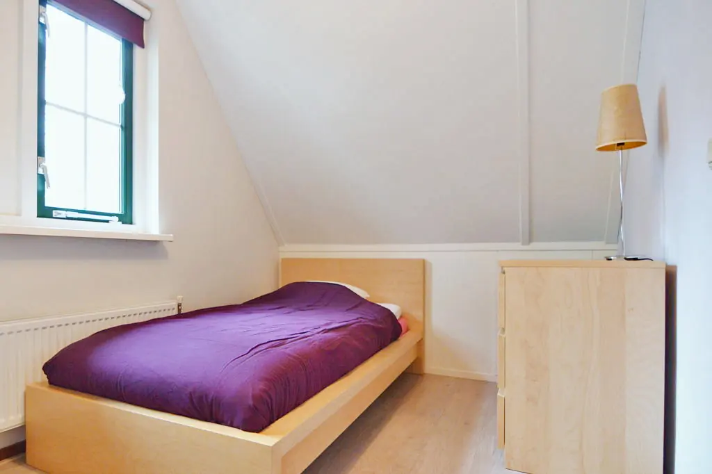 Slaapkamer onder een schuin dak met een eenpersoonsbed, een paars dekbed, een raam met rolgordijn, een radiator en een ladekast met een schemerlamp.