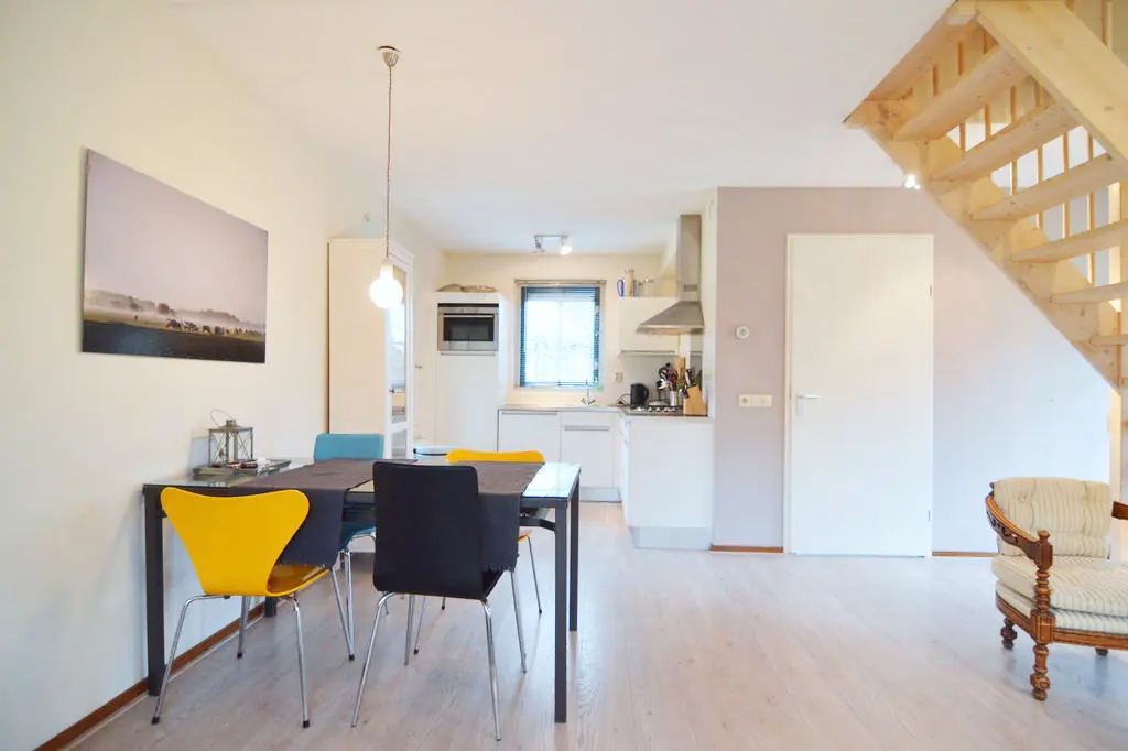 Woon-eetkamer met een eettafel met vier stoelen en een hanglamp, een foto op doek aan de wand, een open houten trap, een witte binnendeur, een fauteuil in de hoek en de keuken op de achtergrond.