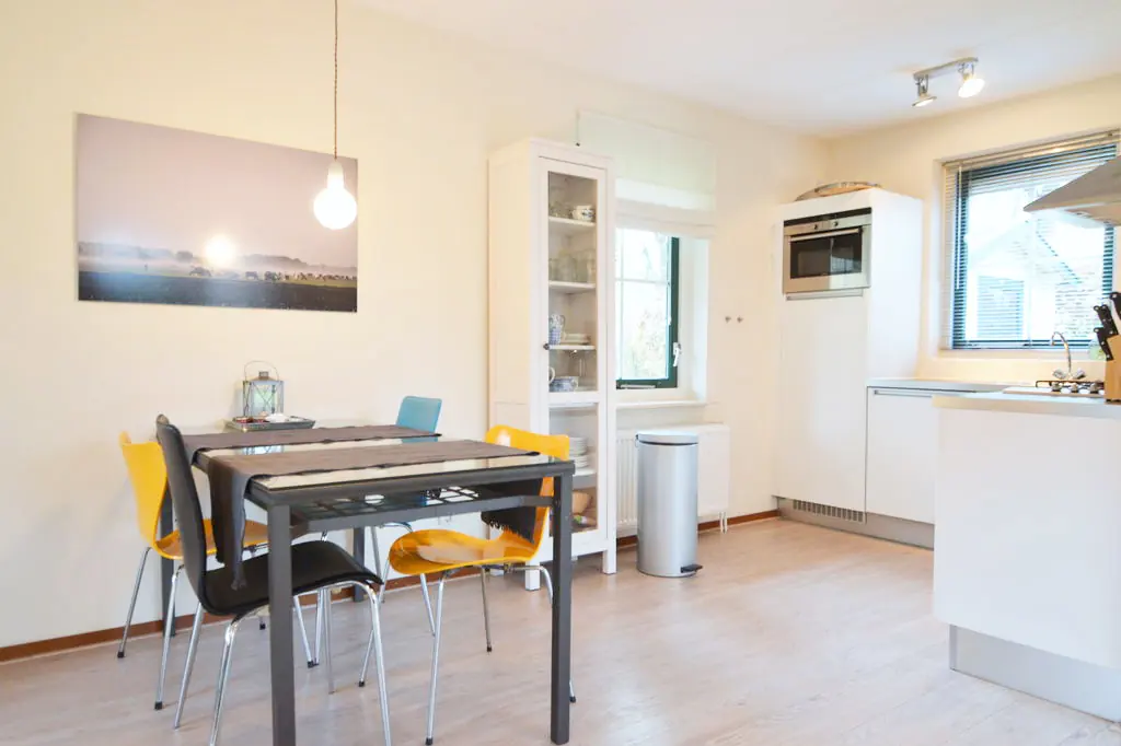 Eettafel met vier stoelen en een hanglamp, een foto op doek aan de wand, een witte vitrinekast met serviesgoed, een pedaalemmer en een radiator onder het raam, met rechts de keuken.