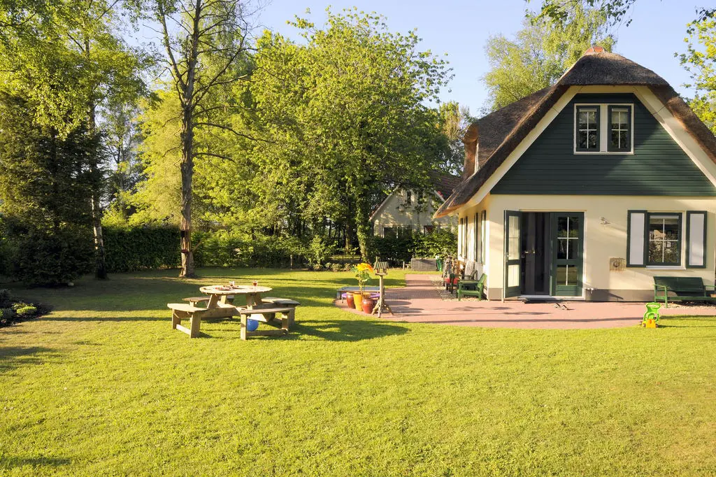 Het vakantiehuis met rieten dak, groene gevel en groene luiken, met openstaande terrasdeuren, een bakstenen terras, een picknicktafel en een gazon ervoor.