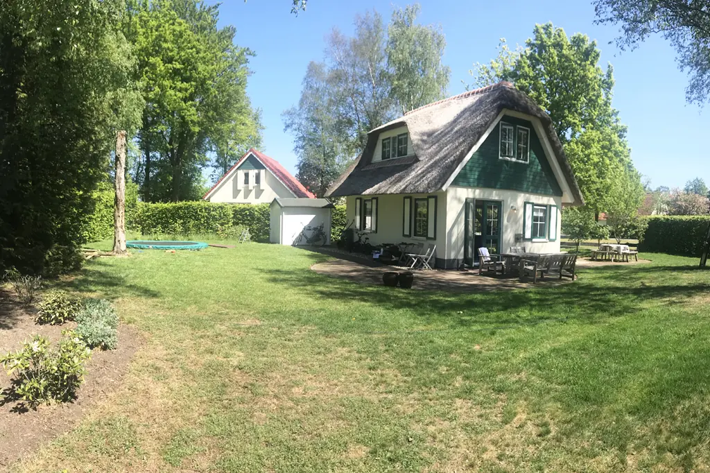 Vrijstaand huis met rieten kap, groene luiken en witte gevel, met een gazon ervoor, een terras met een houten tafel, banken en klapstoelen, fietsen tegen de muur, een schuurtje met witte deur en een verzonken trampoline in het gras.