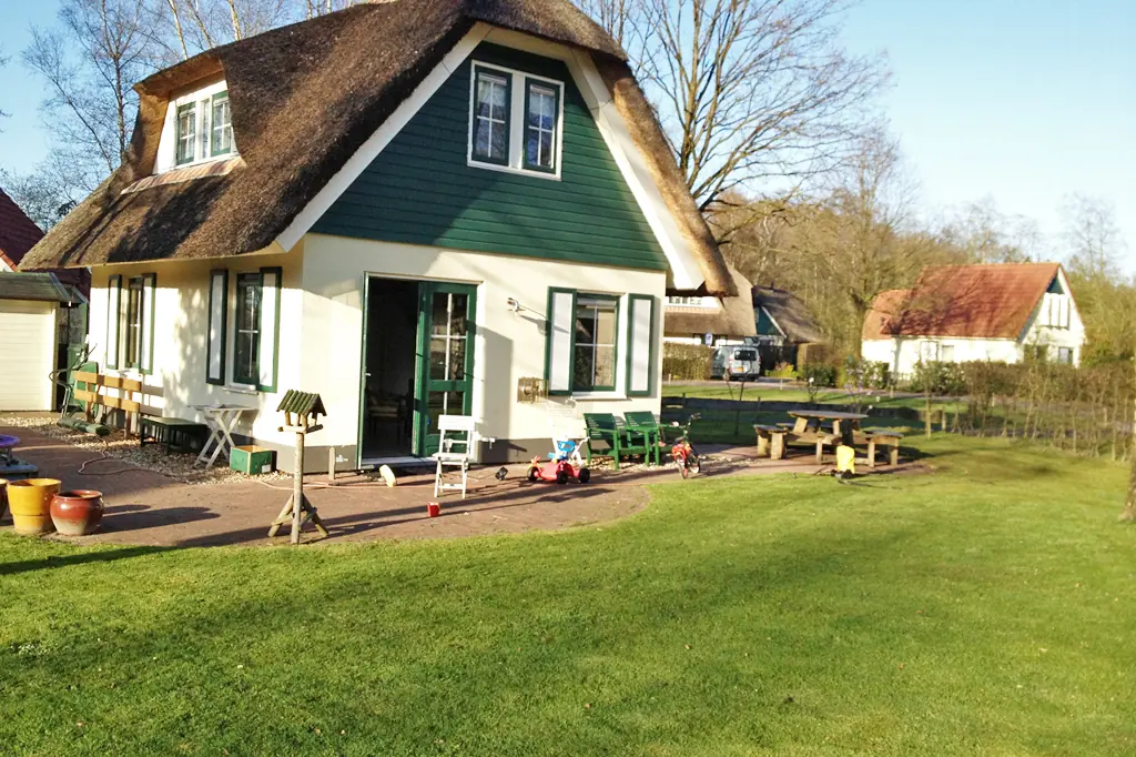 Het vakantiehuis met rieten dak van de zijkant gezien, met een openstaande deur, een terras met tuinstoelen en een vogelhuisje, en een gazon ervoor, met kale bomen.