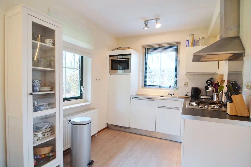 Keuken met witte kasten, een gasfornuis onder een roestvrijstalen afzuigkap, een inbouwoven in een hoge kast, een spoelbak met kraan onder het raam, een vitrinekast met glazen, mokken en borden, een pedaalemmer en een radiator onder het raam.