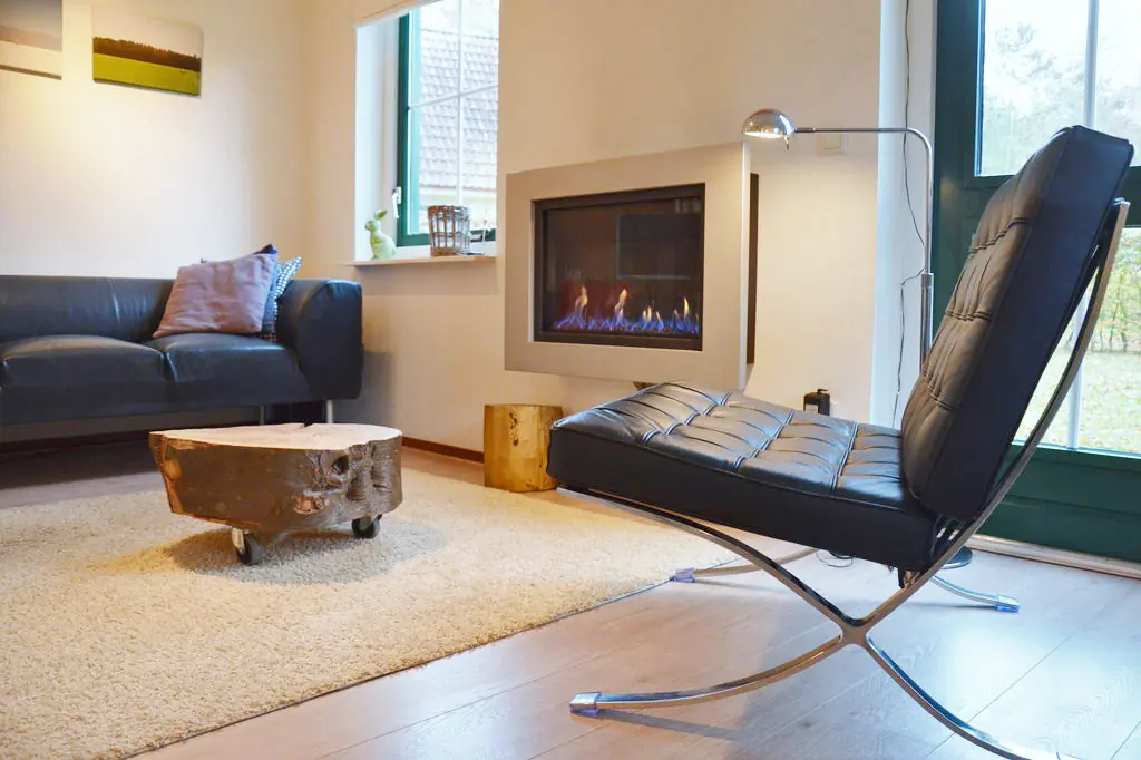 Woonkamer met een donkere leren bank, een ronde salontafel van een boomstam op wielen, een ingebouwde gashaard, een zwarte leren fauteuil op een chromen frame en ramen met uitzicht op gras en bomen.