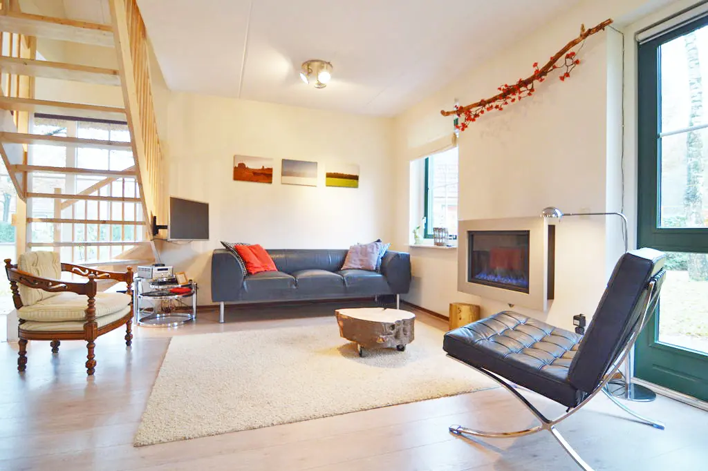 Woonkamer met een grijze leren bank, een zwarte leren stoel met chromen frame, een salontafel van een boomstamschijf op wielen, een hoogpolig kleed, een ingebouwde gashaard, een televisie aan de muur en een open houten trap langs een groot raam.