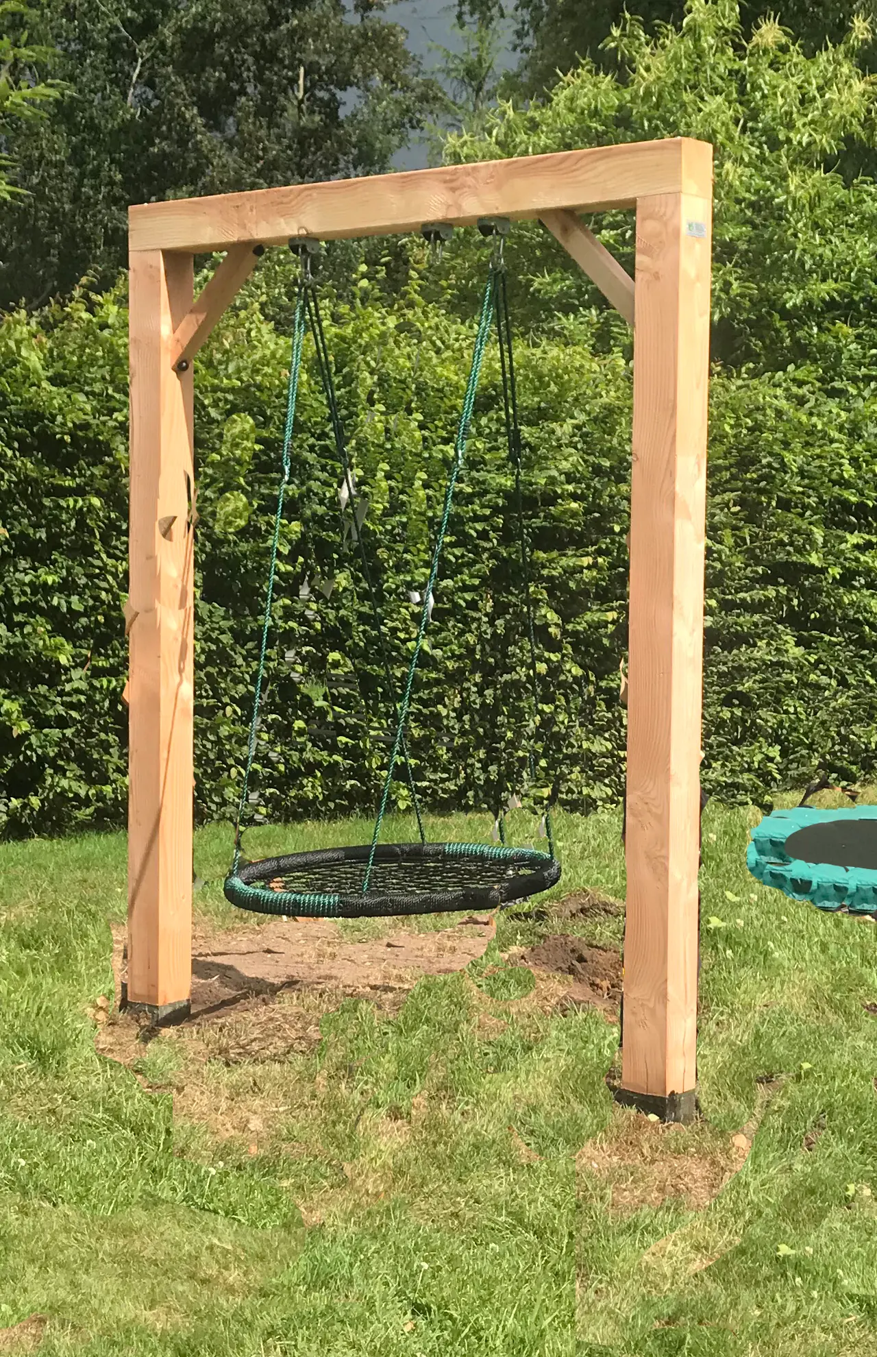 Een houten schommelframe met een ronde nestschommel in het gras, met een ingegraven trampoline ernaast en een beukenhaag erachter.