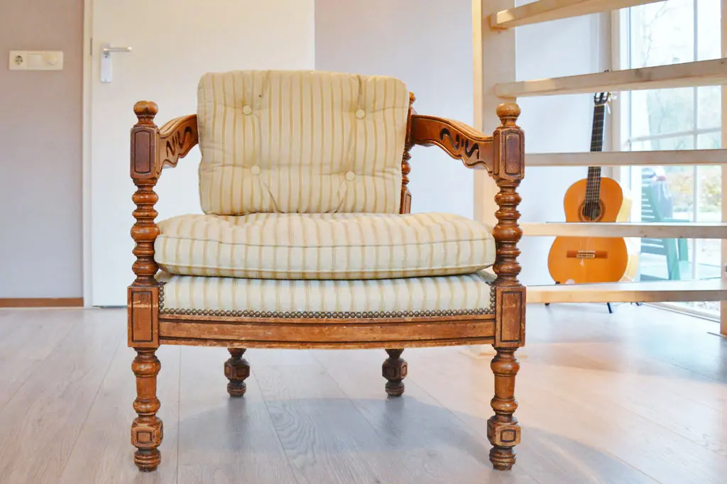 Houten fauteuil met gestreepte kussens op een lichte houten vloer, met daarachter een open houten frame, een klassieke gitaar tegen het raam en groene tuinstoelen buiten.